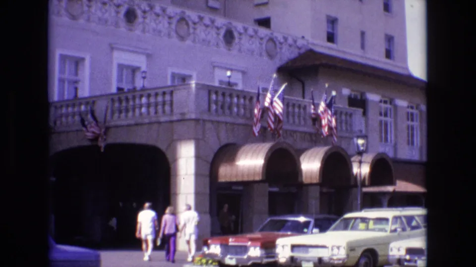 A 1974 Colorado Springs street view shows three pedestrians nearing a shaded arched building entrance while American flags hang above parked mid-century cars.