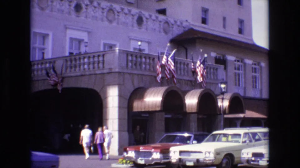The home-movie frame shows three adults in light and purple clothing walking along the curb toward an ornate Colorado Springs building frontage with flags and parked cars.