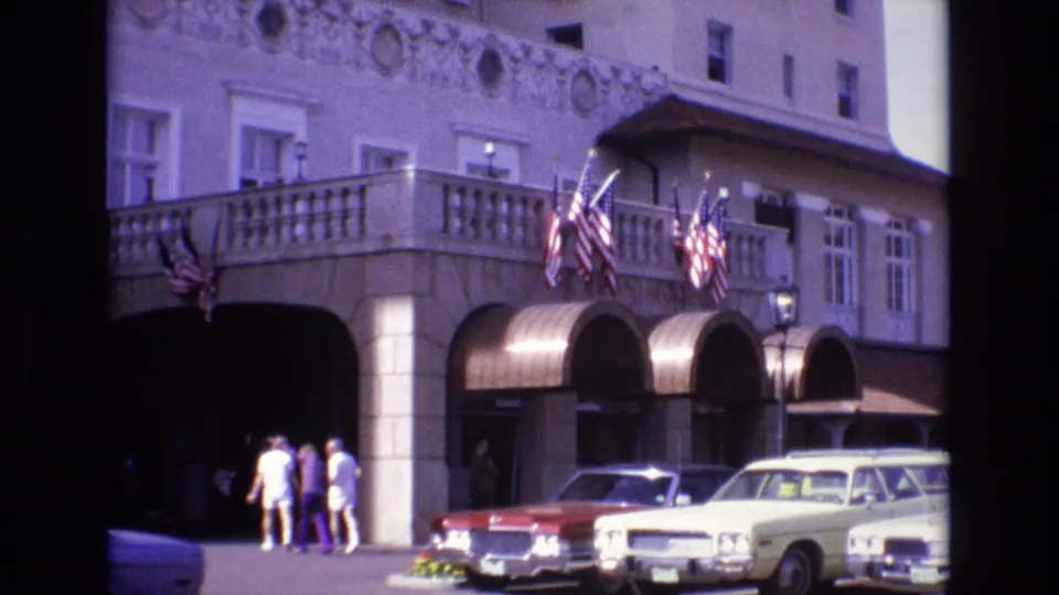 At the end of the clip, three pedestrians are near the shaded arched entrance of the ornate Colorado Springs building, with American flags and 1970s cars visible in front.