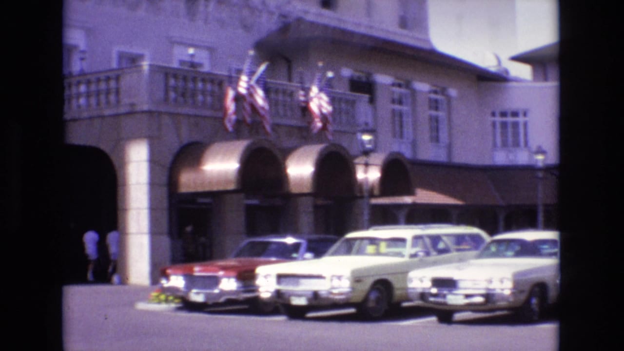 1974 8mm frame from Colorado Springs showing a hotel frontage with arched awnings, American flags, a balcony, and parked sedans and station wagons.
