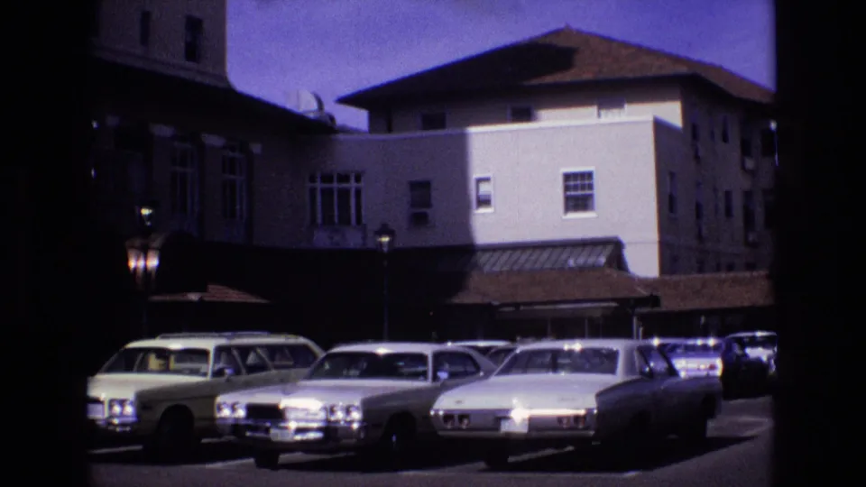 1974 8mm frame in Colorado Springs showing a row of parked cars beneath a hotel facade, windows, roofline, and shaded entrance area.