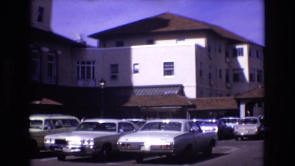 1974 Colorado Springs frame showing a hotel exterior and parking lot with multiple parked sedans, a station wagon, windows, rooflines, and strong afternoon light.