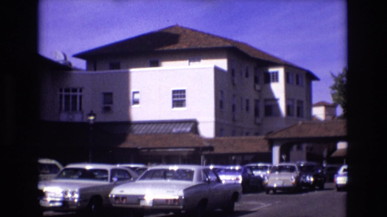 White building with brown roof behind parked 1970s cars in a paved lot.