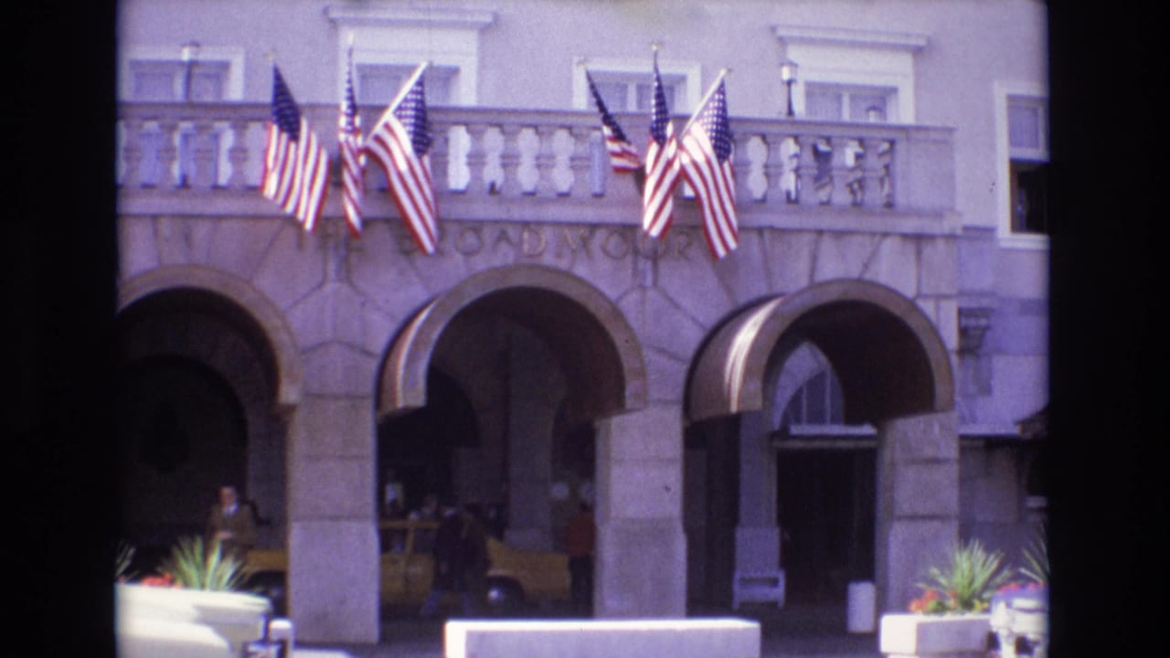 1974 Colorado Springs home-movie frame showing The Broadmoor entrance with American flags, arched stone doorways, planters, people, and a yellow taxi.