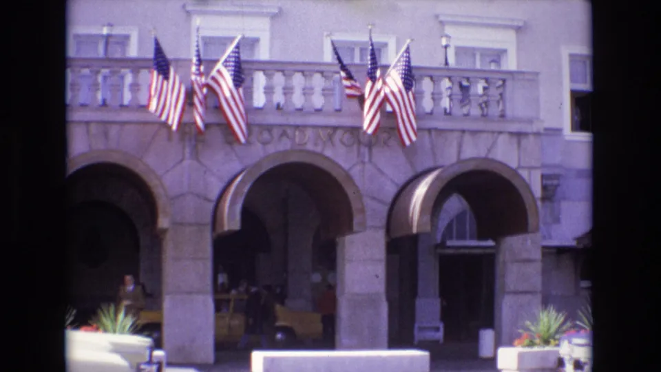 1974 Colorado Springs home-movie frame showing The Broadmoor entrance with American flags, arched stone doorways, planters, people, and a yellow taxi.