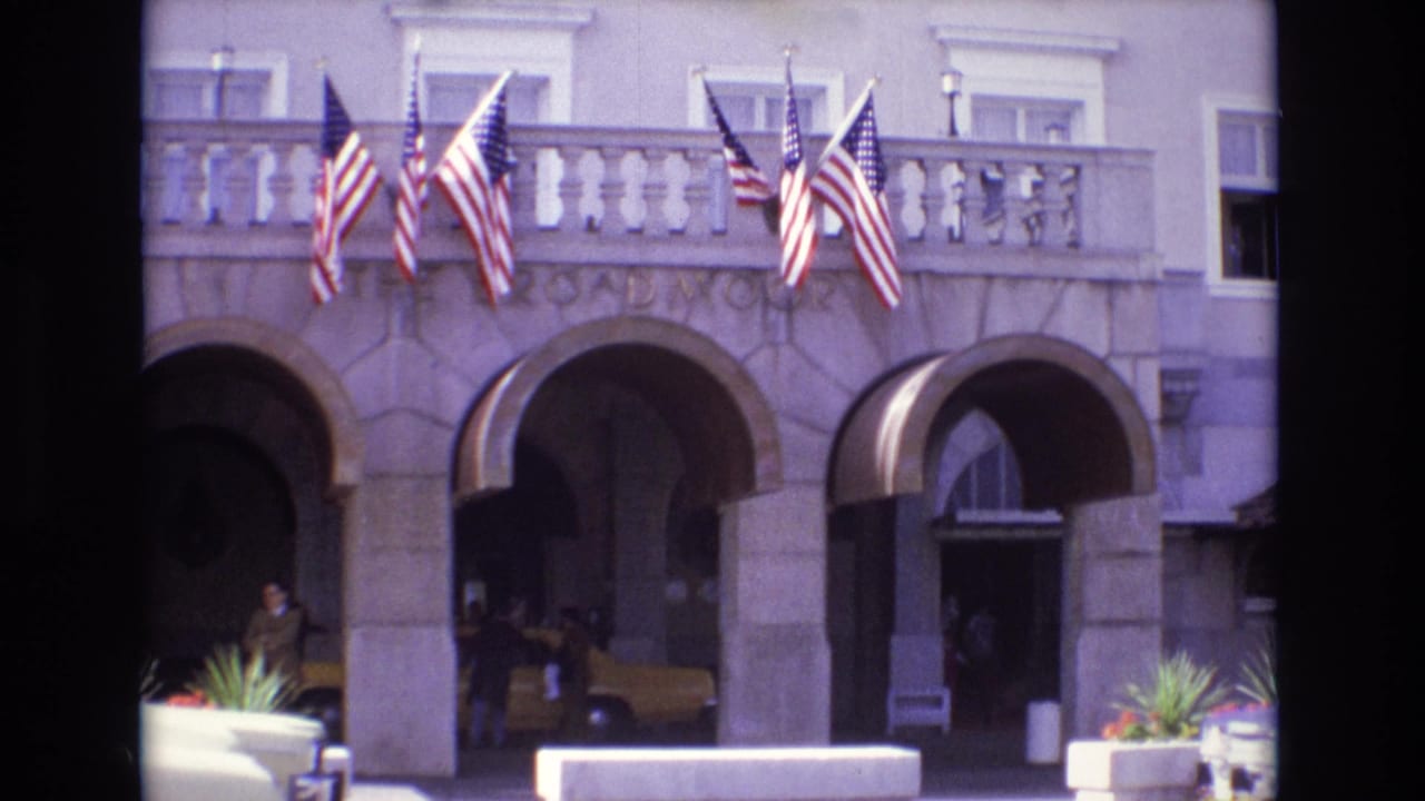 1974 Colorado Springs frame of The Broadmoor hotel entrance with flags on the balcony, arched openings, a yellow taxi, and people near the curb.