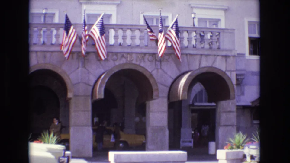 1974 Colorado Springs home-movie still showing the Broadmoor entrance facade, American flags, arched stone bays, planters, people, and a yellow taxi partly visible.