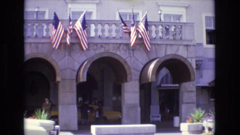 1974 Colorado Springs frame showing The Broadmoor entrance from across the curb, with American flags above the arches, people near the doorway, and a yellow taxi in shadow.