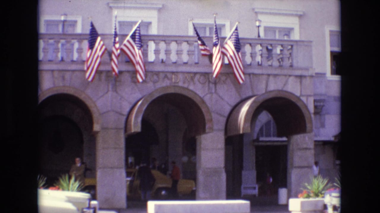 1974 Colorado Springs resort hotel frontage with multiple American flags above arched stone entrances and people near a yellow taxi.