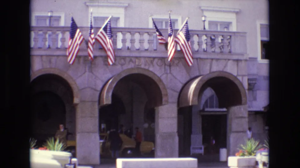 1974 Colorado Springs resort hotel frontage with multiple American flags above arched stone entrances and people near a yellow taxi.