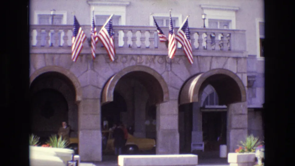 1974 Colorado Springs hotel entrance seen from a visitor's 8mm camera, with American flags, arched stonework, a taxi, and people by the curb.