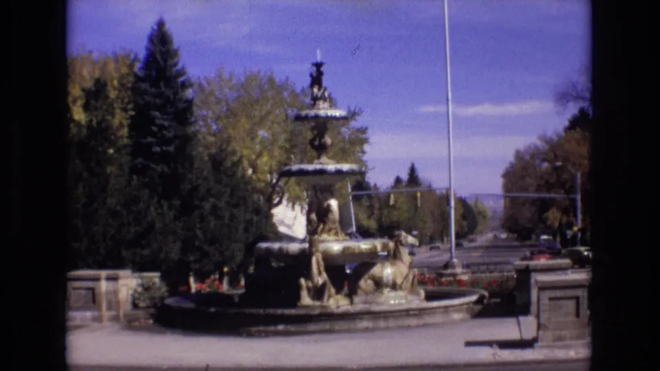 1974 Colorado Springs outdoor fountain scene with a multi-tiered sculptural fountain, surrounding trees, stone edging, and clear blue sky.