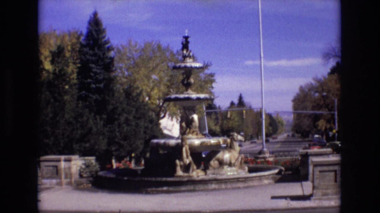 1974 Colorado Springs home-movie frame showing a sculptural multi-tiered fountain with water, evergreen trees, flower beds, stone posts, and a flagpole under a blue sky.