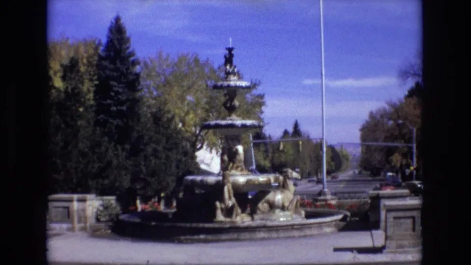 Final 1974 Colorado Springs frame of the fountain scene, with the multi-tiered water feature, flagpole, flower beds, trees, stone posts, and distant roadway held in soft 8mm color.