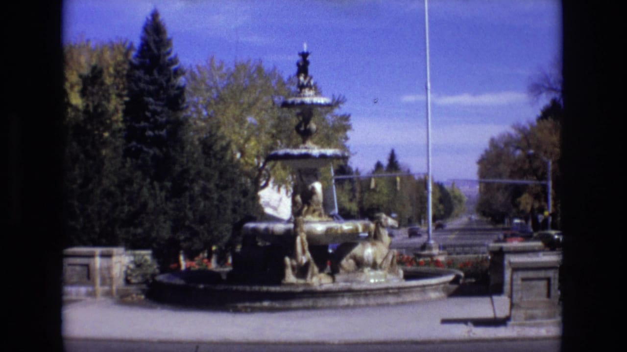 1974 Colorado Springs view of the fountain and landscaped median, with trees surrounding the stone basin and cars visible on the roadway beyond.