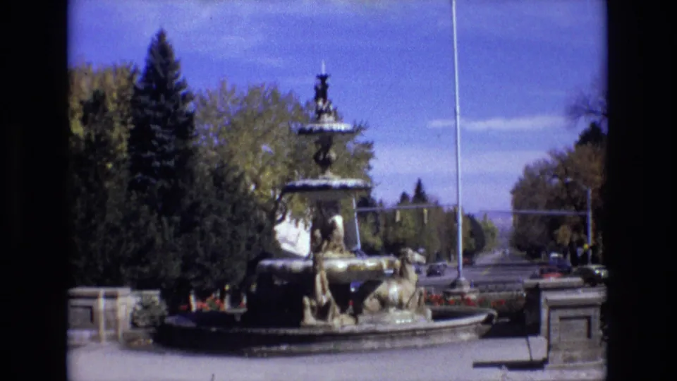 Wide 8mm frame of the Colorado Springs fountain from across the street, showing sculpted figures, a stone basin, trees, road lanes, and distant vehicles.