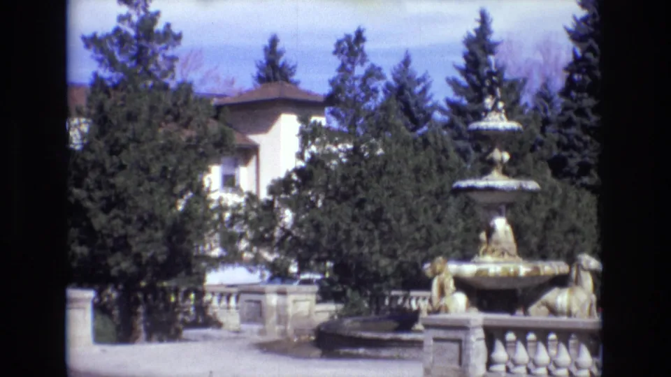 Side angle of the Colorado Springs fountain in 1974, with sculpted figures, stone railing, dense trees, and a large light-colored building partly visible behind foliage.