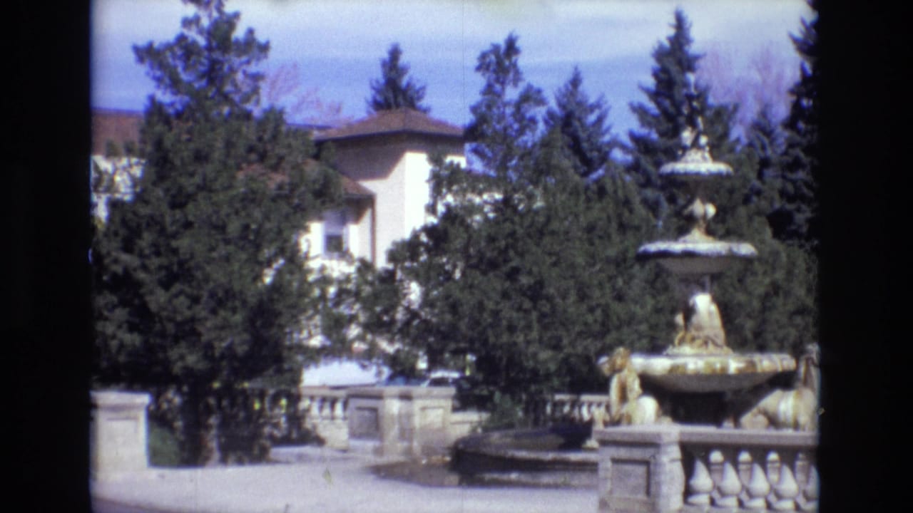 1974 Colorado Springs home-movie frame showing a multi-tiered stone fountain with lion figures, a balustrade, dense trees, and a large light-colored estate building beyond.