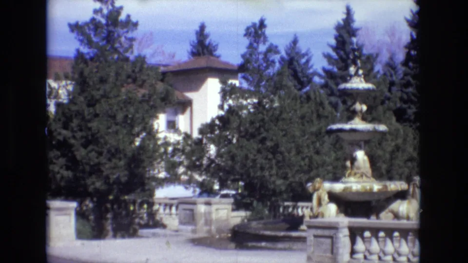 1974 Colorado Springs home-movie frame showing a multi-tiered stone fountain with lion figures, a balustrade, dense trees, and a large light-colored estate building beyond.