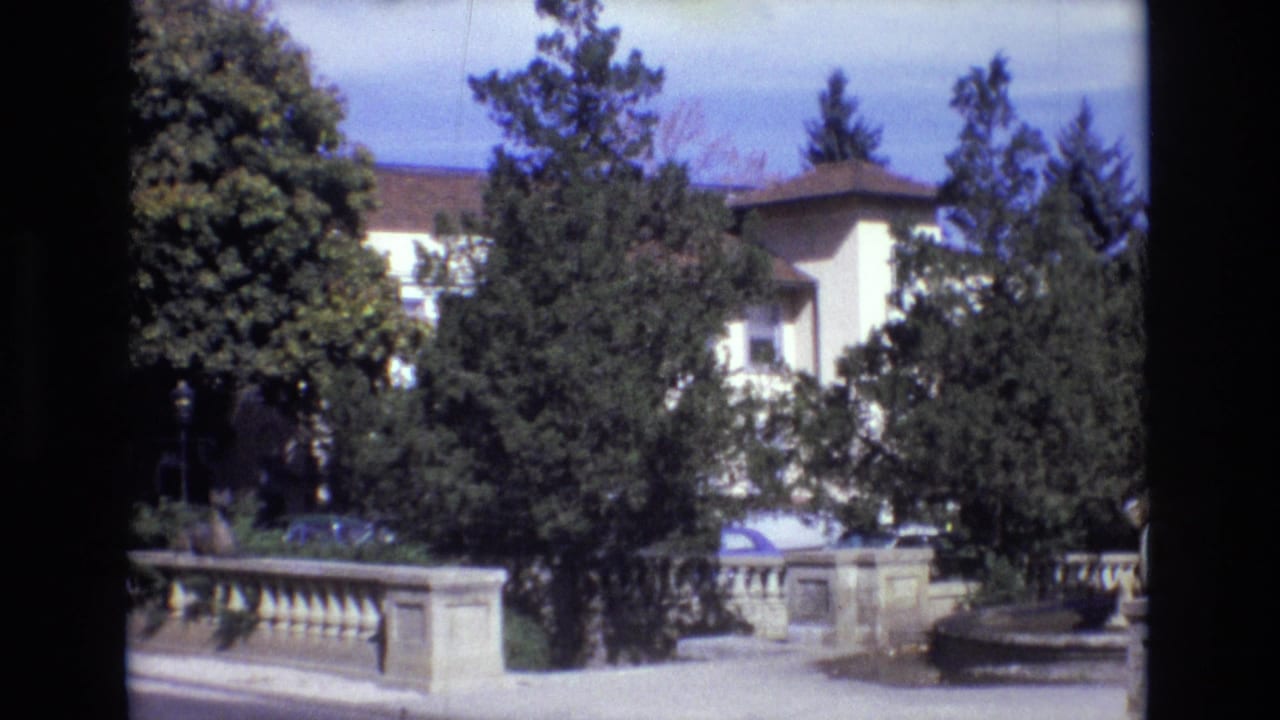 1974 Colorado Springs home-movie frame showing a stone balustrade, dense landscaping, a partly visible parked car, and the estate building behind the trees.