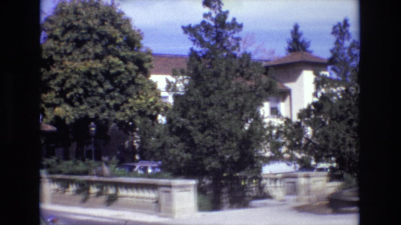 1974 Colorado Springs estate exterior with a pale mansion-like building behind trees, a stone balustrade along the drive, a parked car, and the fountain cropped at the right edge.