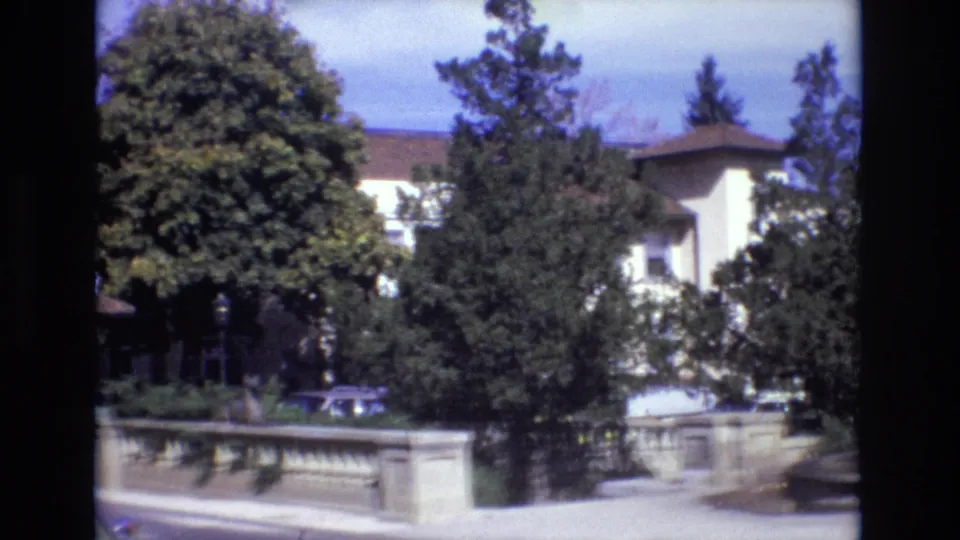 1974 Colorado Springs estate exterior with a pale mansion-like building behind trees, a stone balustrade along the drive, a parked car, and the fountain cropped at the right edge.