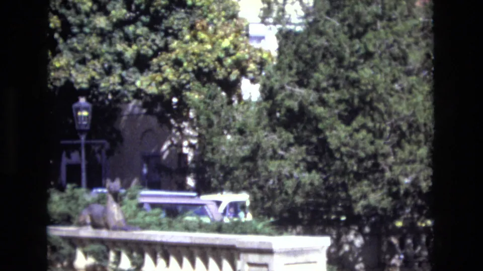1974 Colorado Springs home-movie frame with a small decorative statue on or near a stone balustrade, dense greenery, a lamppost, and a car partly hidden below the railing.