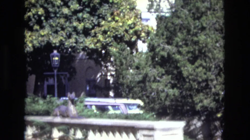 1974 Colorado Springs frame showing a sunlit stone balustrade with a small decorative statue, leafy trees, a lamppost, and a light-colored car partly obscured by the railing.