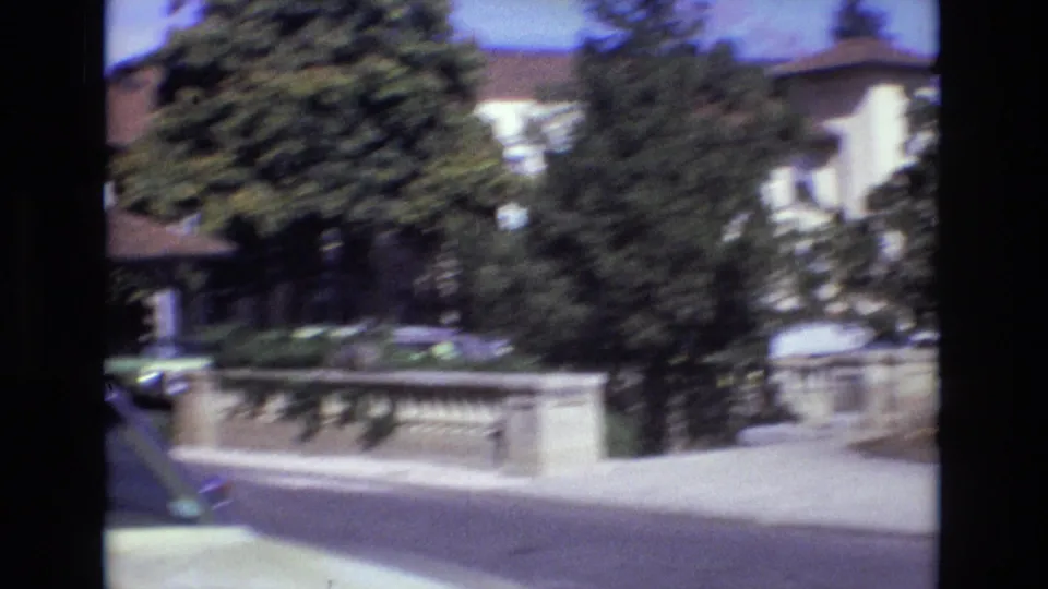 Handheld 1974 Colorado Springs frame showing motion blur as the camera passes a stone railing, trees, road, and light-colored buildings.
