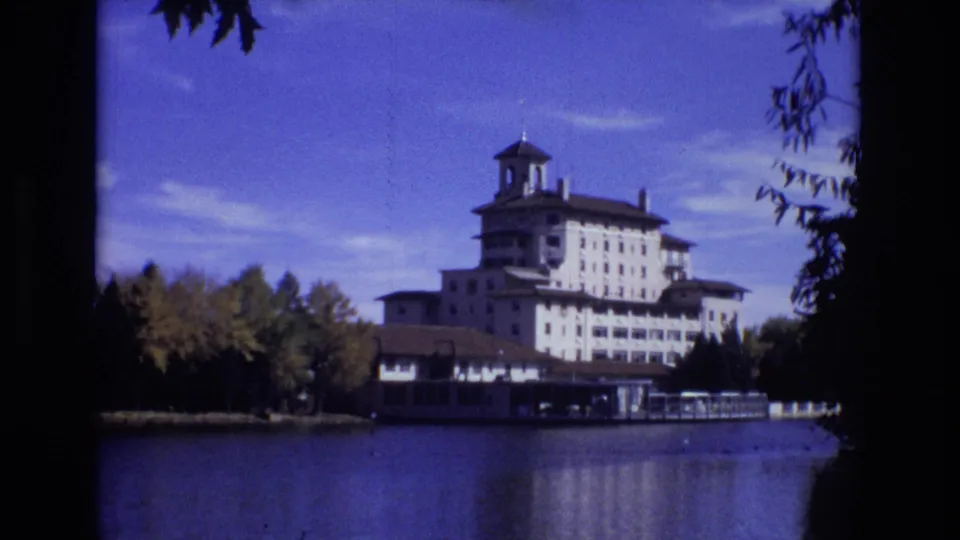 Wide 1974 Colorado Springs view of the Broadmoor Hotel across a calm lake, with shoreline trees and reflections under a clear blue sky.