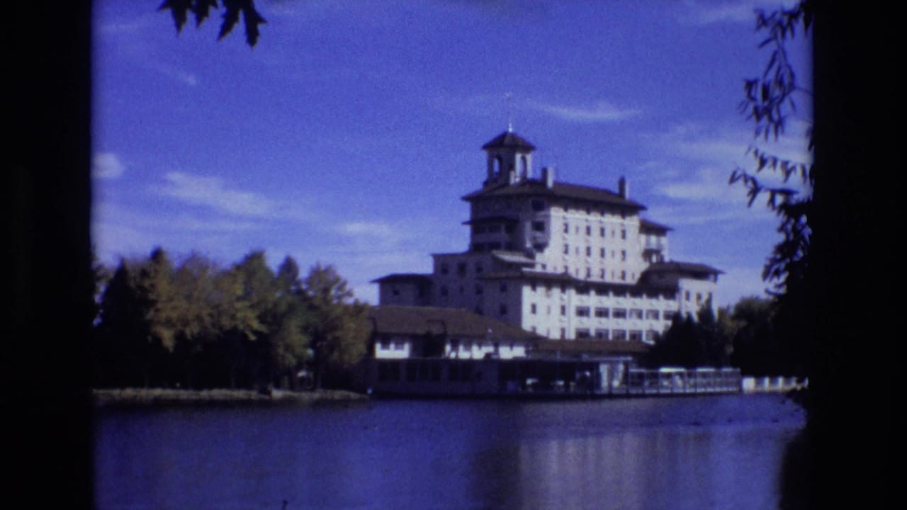 1974 8mm view of the Broadmoor Hotel in Colorado Springs across calm lake water, framed by shoreline trees and dark leaves in the foreground.
