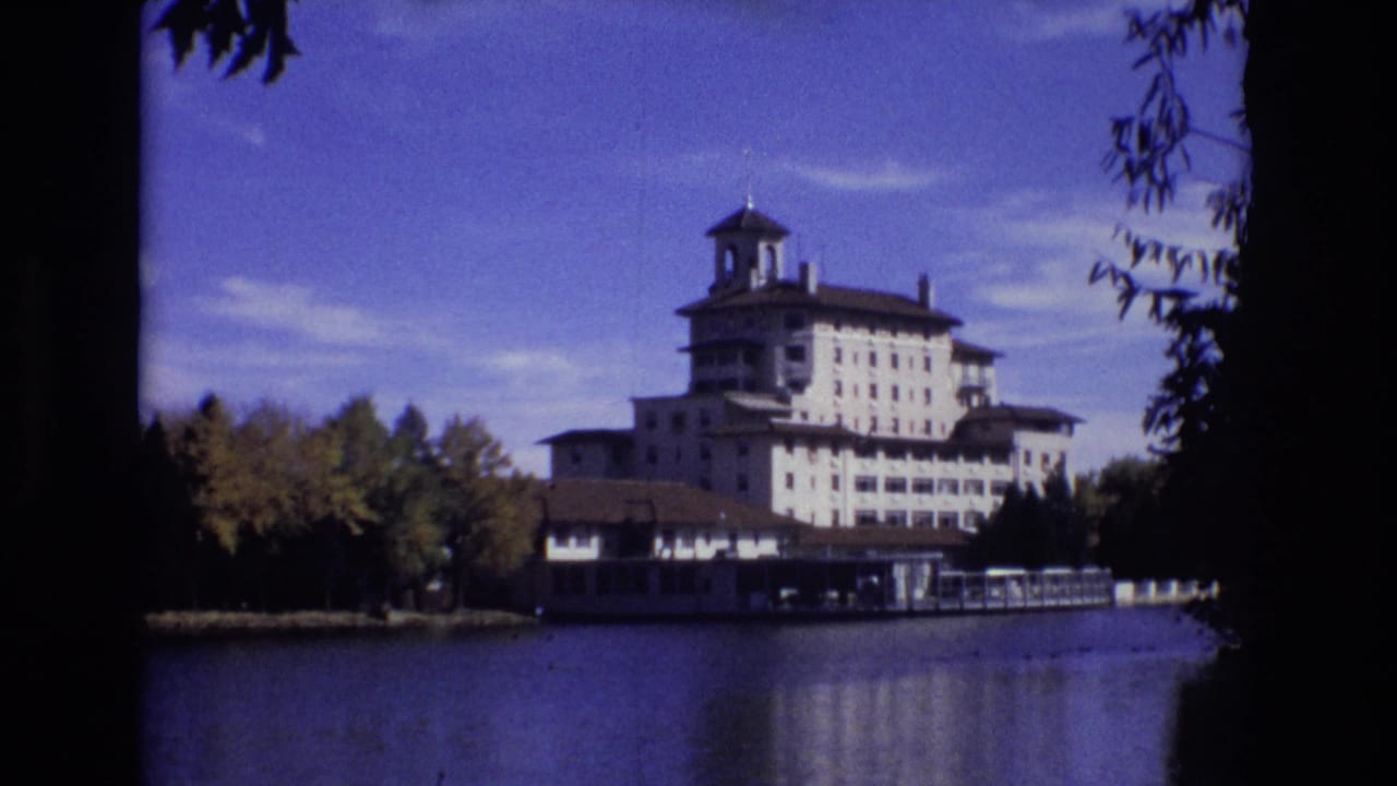 White lakeside hotel with bell tower seen across a calm lake.