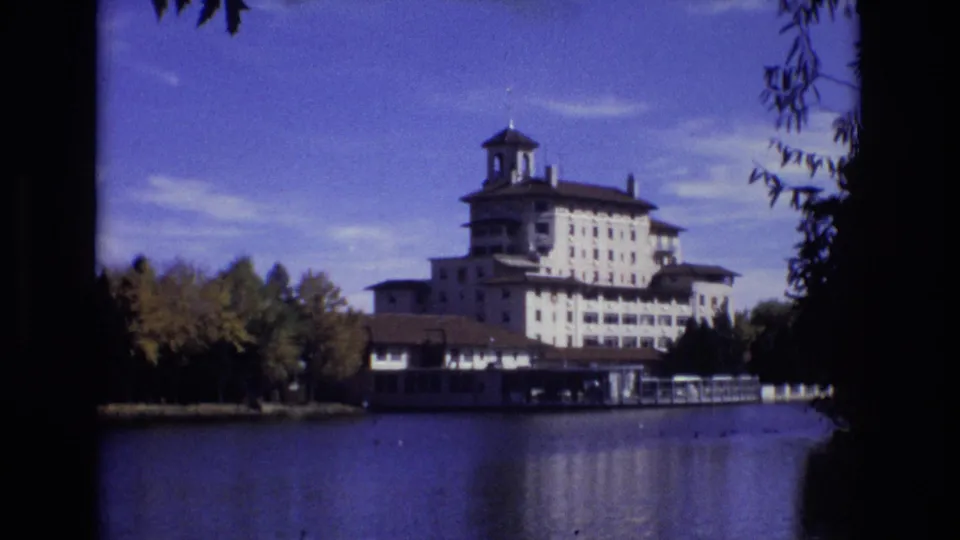 A steady 1974 8mm view holds on the Broadmoor Hotel above the lake, with autumn trees along the shore and dark foliage framing the right edge.