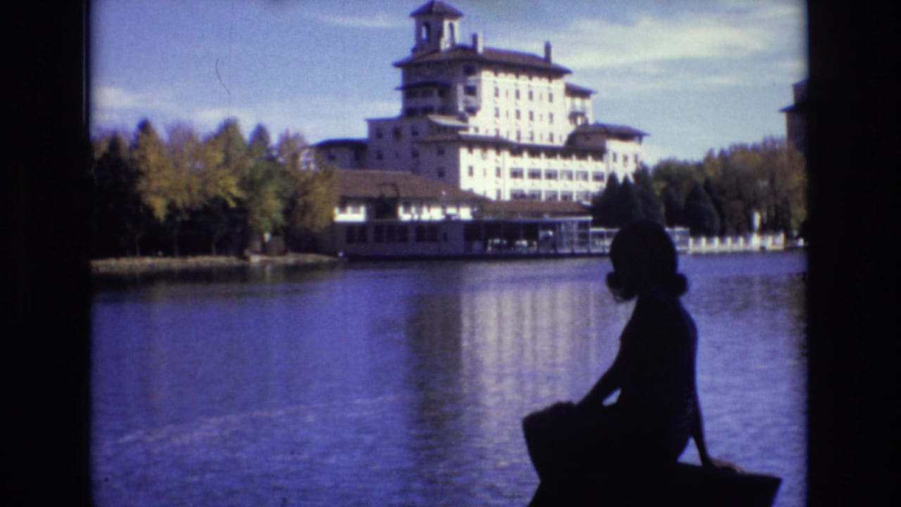 1974 Colorado Springs home-movie frame showing a woman in dark silhouette seated near calm lake water, facing a large ornate hotel with a tower and autumn trees along the shore.