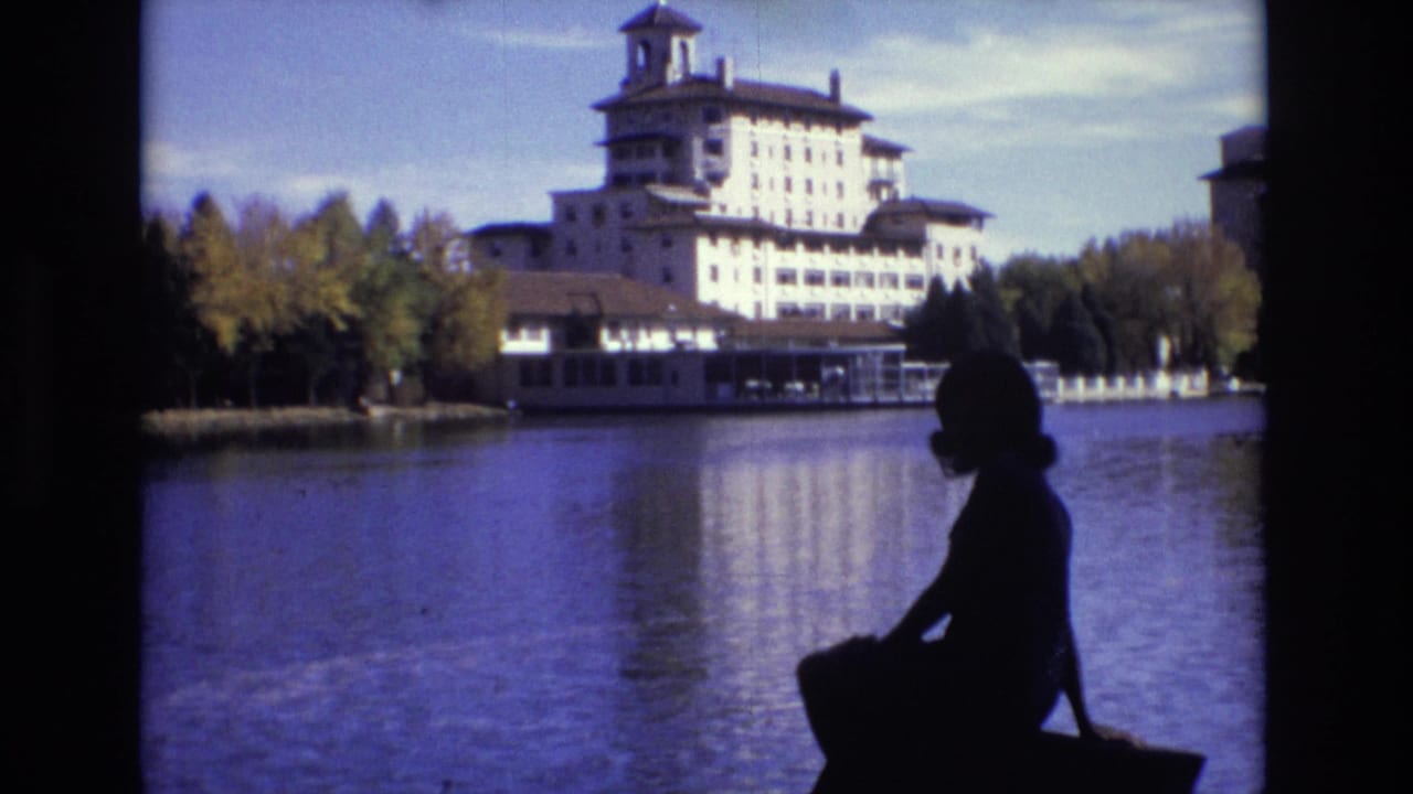 1974 Colorado Springs frame with a seated woman in silhouette in the foreground, calm water in the middle ground, and a large towered hotel backed by trees across the lake.
