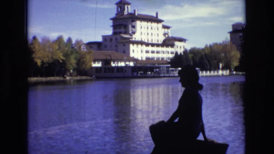 1974 Colorado Springs frame showing a seated woman in profile-like silhouette at right, looking across calm lake water toward a large towered hotel and tree-lined shore.