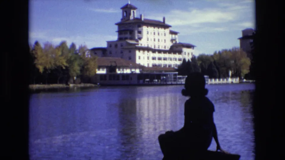 1974 8mm Colorado Springs frame showing the woman lower in the foreground, with the Broadmoor Hotel, trees, and lake reflections in the background.