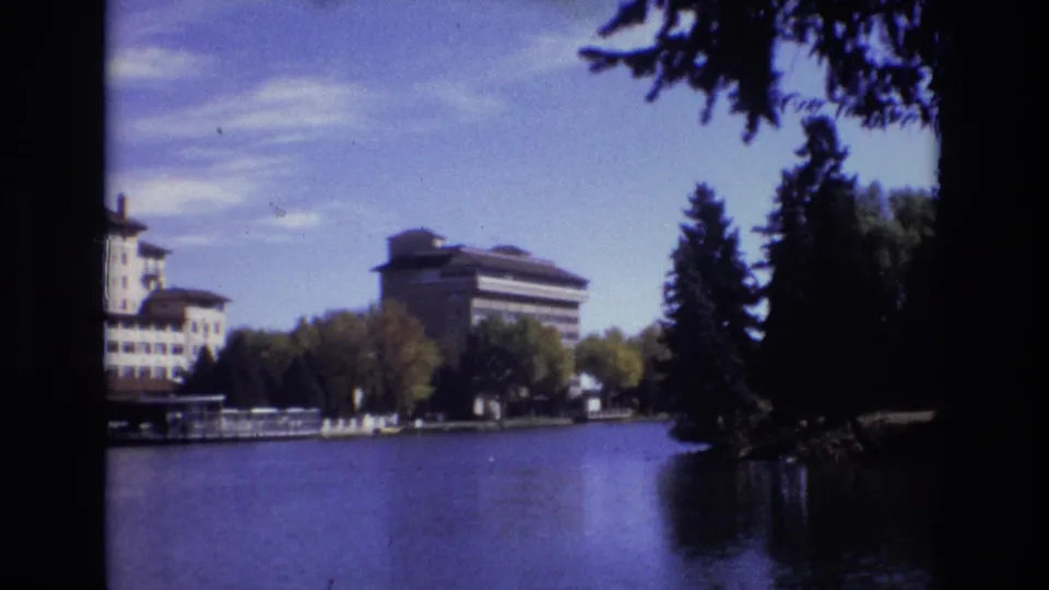 1974 Colorado Springs 8mm frame showing a wide calm lake, far-shore trees, evergreen silhouettes, and resort-style buildings partly visible at left and center.