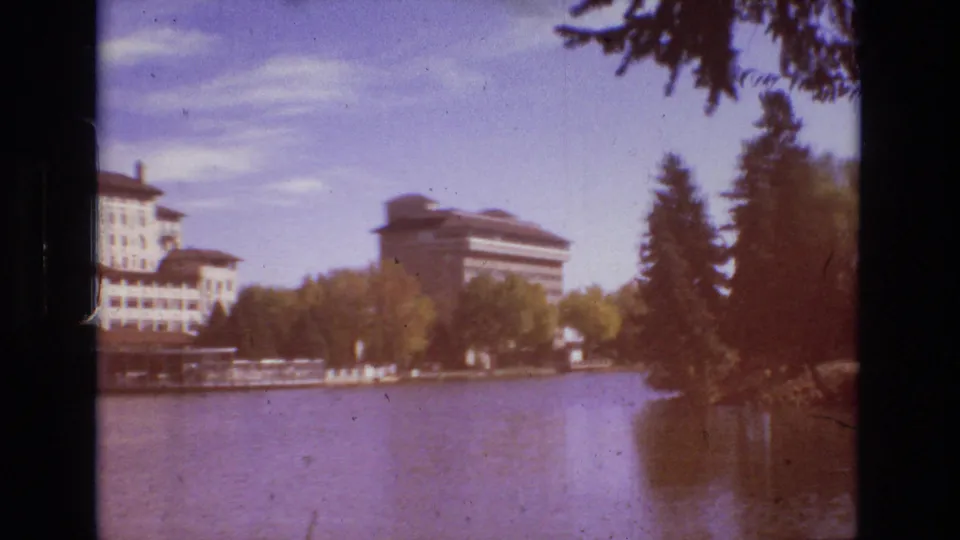 1974 Colorado Springs 8mm frame with a warm color shift, showing calm lake water, large buildings beyond the shore, trees, and a reddish flare near the right edge.