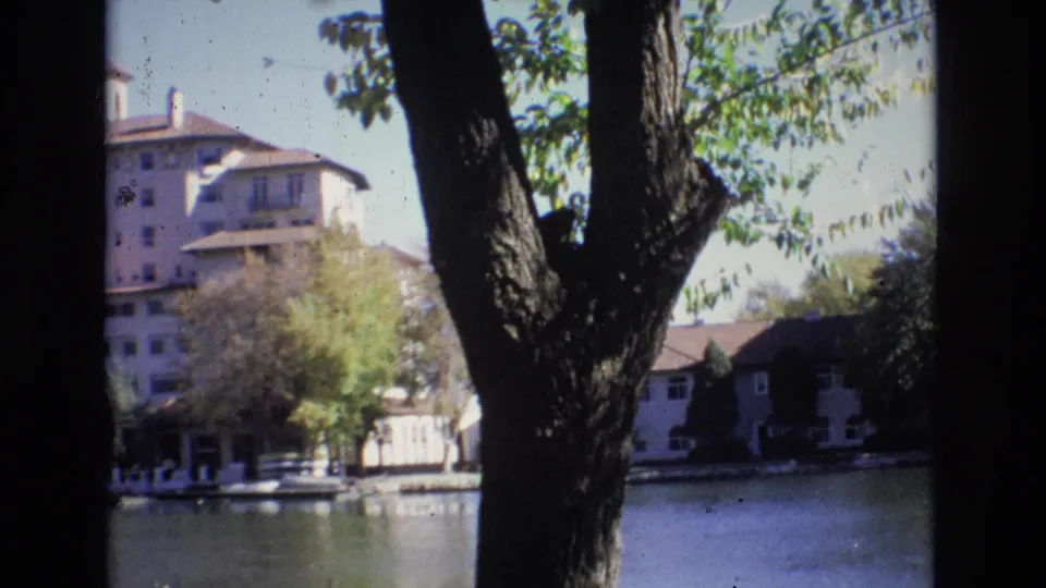 The frame shows a large foreground tree, leafy branches, calm lake water, and hotel-like buildings in a 1974 Colorado Springs 8mm home-movie view.
