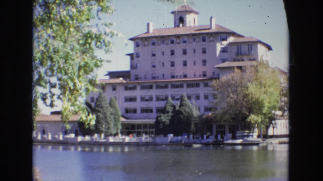 1974 8mm home-movie view of the Broadmoor Hotel in Colorado Springs seen across calm water, with trees framing the resort building and distant visitors along the shoreline.