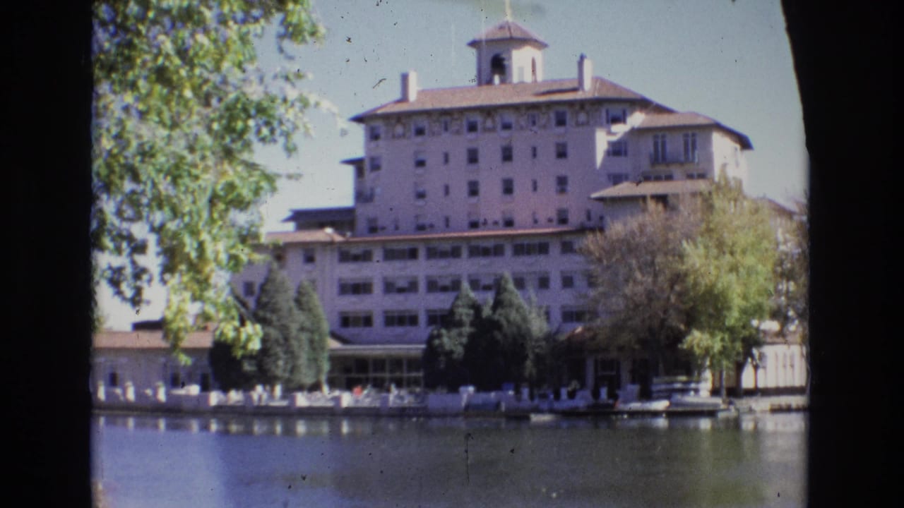 1974 Colorado Springs view of the Broadmoor Hotel from across the lake, showing the white facade, roof cupola, trees, and small distant people near the waterline.