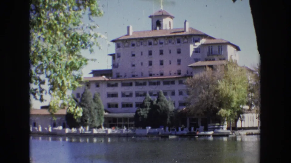 Home-movie frame of the Broadmoor Hotel in 1974, with leafy branches at left, the lakeside building centered, and faint scratches and grain visible across the scan.