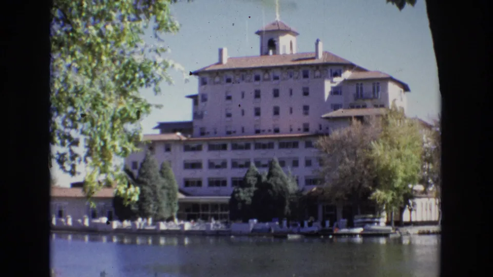 1974 8mm view of the Broadmoor Hotel across the lake in Colorado Springs, with the building, cupola, trees, shoreline activity, and soft water reflections visible.