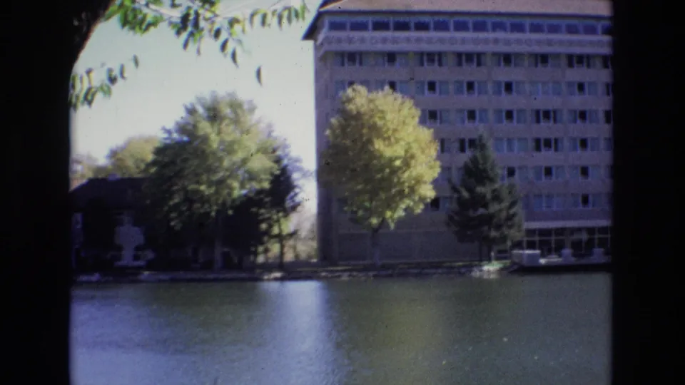 1974 Colorado Springs home-movie frame showing a long hotel wing along the water, with trees in front and a soft reflected surface below.