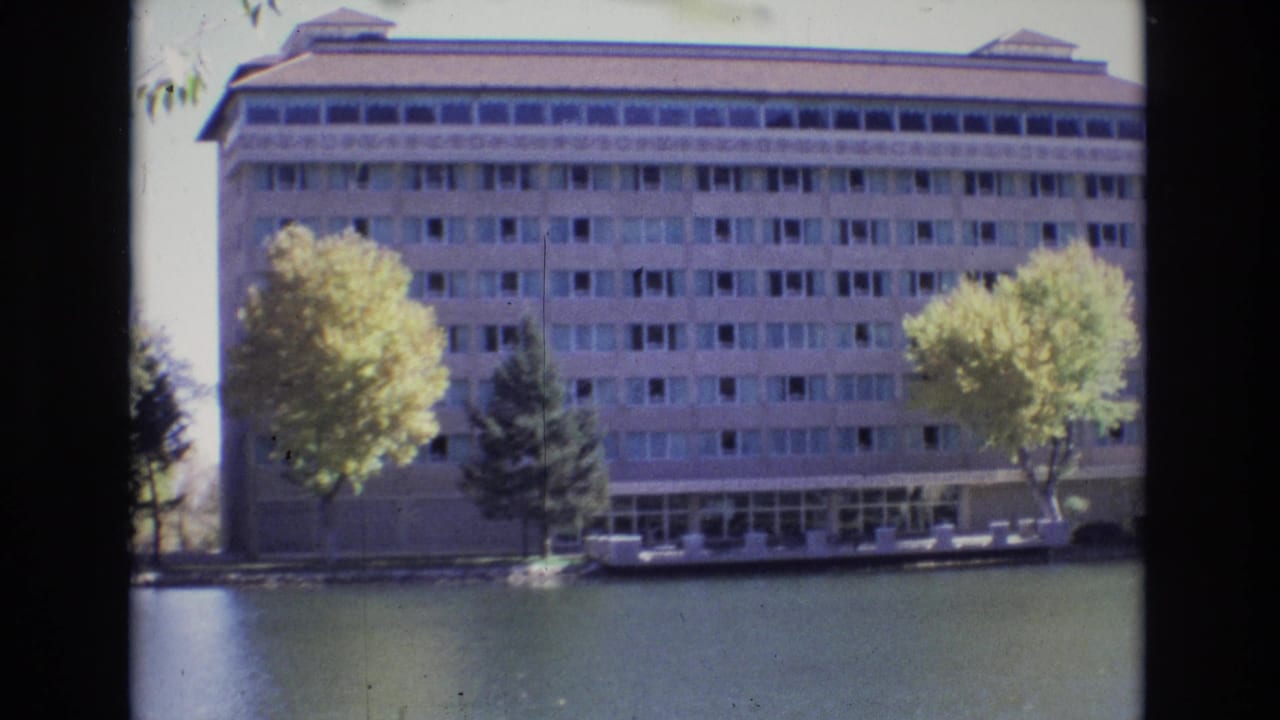 Large multi-story building across calm water with trees in front.
