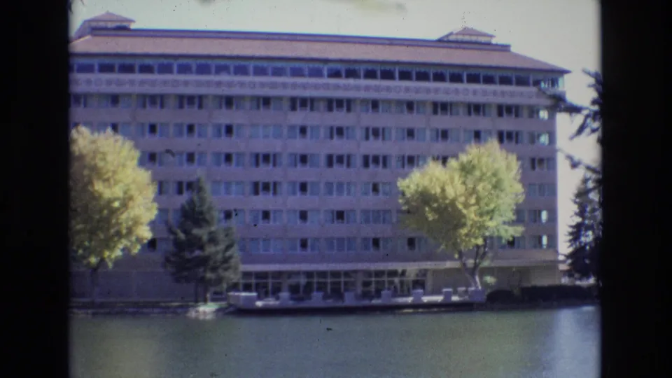 Wide 8mm frame of the Colorado Springs building exterior beside a calm waterway, with repeated window rows, shoreline trees, and a small boat or docked craft visible below.
