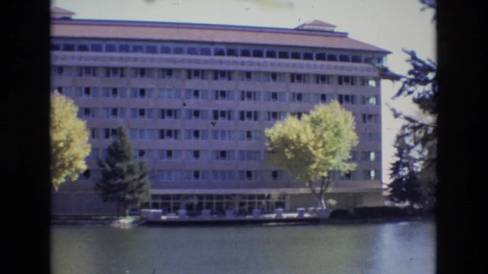 Slightly shifted 1974 frame of the large waterfront building, cropped closer on its windowed facade with autumn trees, an evergreen, and water in the foreground.