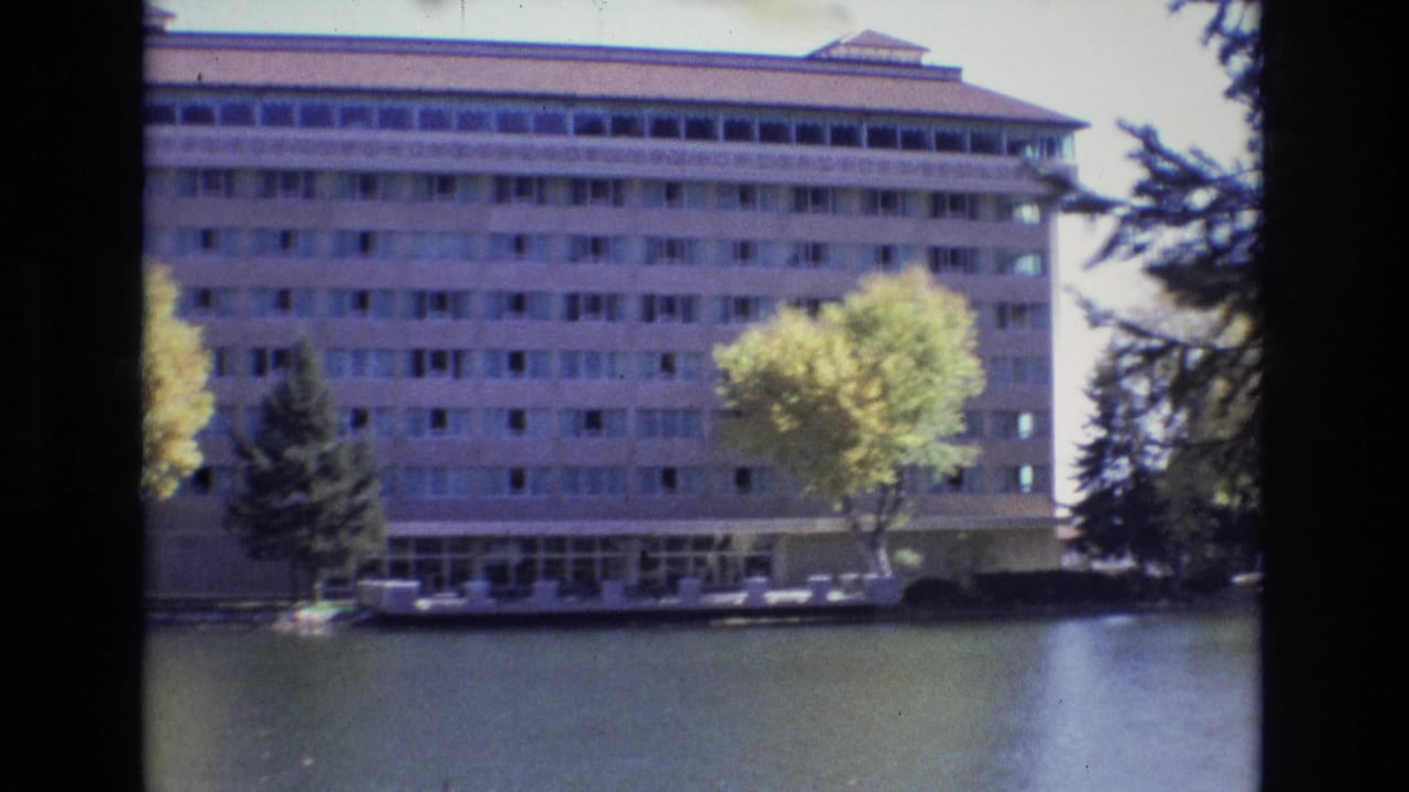 1974 Colorado Springs home-movie frame showing a large mid-century style building across calm water, with a yellow-green autumn tree, evergreens, and a parked car near the ground-floor entrance.