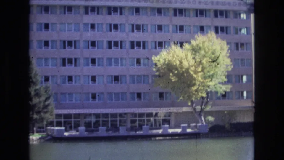 1974 Colorado Springs frame of a broad multi-story building with rows of windows, calm water in front, and a sunlit yellow-green tree partly obscuring the facade.